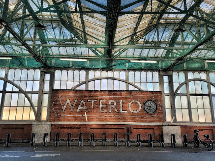 A shot of Waterloo underground station platform showing a white mosaic tiled wall with a prominent London Underground roundel sign displaying 'WATERLOO' in blue on a silver rectangular background. The